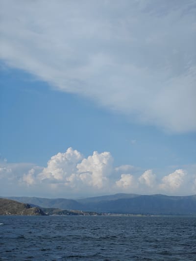 An aerial view captures the deep blue waters of Lake Bukhtarma under a partly cloudy sky, with the distant shores of New Bukhtarma, Kazakhstan, visible. The water exhibits a rough texture with small waves. Along the coastline, a range of green, tree-covered hills and mountains stretch across the horizon. A thin strip of urban development, dotted with buildings, is nestled at the base of the hills. The sky is a light blue, with scattered white, puffy clouds, particularly prominent in the middle ground.