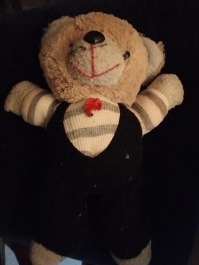 A well-worn, stuffed teddy bear is displayed against a dark background. The bear has a tan, furry head with a black nose and a stitched red smile. Its arms and upper body are made of horizontally striped fabric. It's wearing a black vest and pants. A small, red bow is stitched to its chest. The toy appears to be old and potentially damaged, with visible signs of wear and tear.