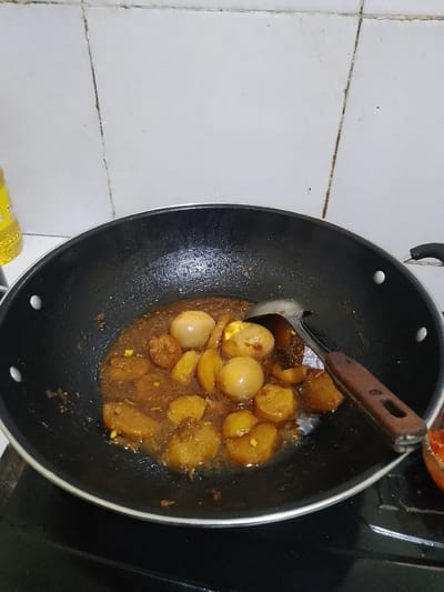 A high-angle medium shot shows a black wok filled with a brown sauce containing several hard-boiled eggs and what appear to be sliced potatoes. A silver ladle with a wooden handle is resting in the wok, partially submerged in the sauce. The wok is placed on a black stovetop. In the background, a white tiled wall with visible grout lines is present. A portion of a yellow bottle, likely oil, is visible on the left side of the frame, and the edge of a red object, possibly a tomato, is seen on the right. The scene appears to be a kitchen, likely in Jonggol, Indonesia.