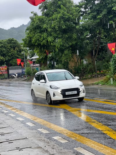 A white Hyundai i10 car drives on a wet, paved road marked with yellow and white lines in Sơn Dương Commune, Vietnam. Rain is falling, creating streaks of water in the air and on the road surface, which reflects the surrounding greenery. The car is moving from left to right in the frame, with its headlights illuminated. In the background, lush green trees and hills are visible. Several red Vietnamese flags with yellow stars are strung up along the side of the road, attached to poles. A parked white scooter and a blue truck are visible on the left side of the road, along with a sign that appears to be advertising furniture. The sky is overcast and gray, indicating continued inclement weather.