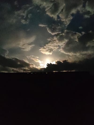 The image depicts a night sky with dramatic clouds illuminated by moonlight. The lower portion of the image is dominated by a dark silhouette of a rooftop, possibly of a building in Қарғалы, Kazakhstan. The clouds are varied, with some appearing bright and wispy, catching the moonlight, while others are dark and heavy, casting shadows. The moon itself is partially obscured by the clouds, creating a luminous halo effect and highlighting the texture and movement of the atmospheric formations. The overall impression is one of a moody and dynamic night sky over a dark, unidentifiable urban or suburban landscape. There are no people or discernible actions visible.
