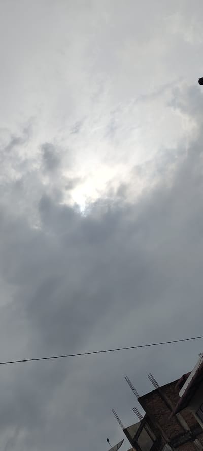 The image shows a cloudy sky with the sun partially obscured by clouds. In the lower right corner, the roof of a building is visible, with exposed rebar suggesting it is under construction. A thin black wire runs horizontally across the middle of the frame. Satellite dishes are also present on the roof. The scene appears to be outdoors in an urban or semi-urban environment in Meunasahceubrek, Indonesia.