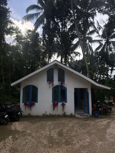 The image shows a white A-frame building with blue-trimmed windows. There are three arched windows with dark interiors and flower boxes containing red flowers. The building also has a rectangular doorway. Palm trees are visible in the background. The ground is dirt and gravel, and some parked motorcycles are present. The building is located in Tegal Suci, Indonesia.