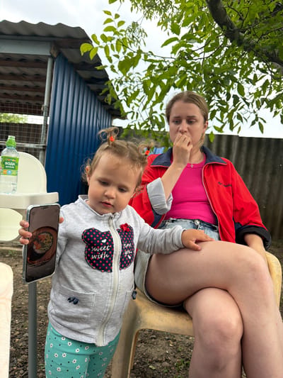 In an outdoor, rural setting, likely in Komsomolskiy, Russia, a young child with blonde hair styled in two ponytails is holding up a smartphone. The child is wearing a grey zippered jacket with red and blue accents and a pink heart patch that says "sweet with love," and light blue pants with a white floral pattern. The child is focused on the phone's screen, which displays an image of sushi. To the right, an adult woman with blonde hair sits with her legs crossed, wearing a bright pink top and a red jacket with blue trim. She is looking downwards with her hand near her mouth, appearing contemplative or concerned. Behind them, a blue corrugated metal structure, a white plastic chair, a bottle of water, and some greenery from a tree are visible, suggesting a casual outdoor gathering or meal. The ground appears to be dirt with some scattered leaves and debris.