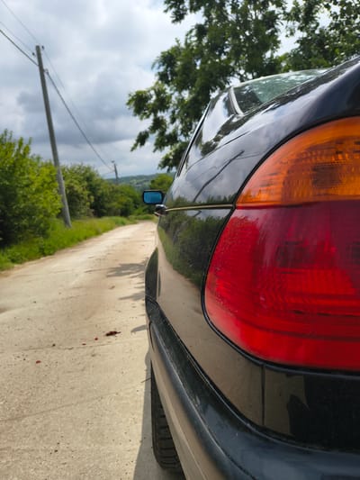 The image displays the rear right side of a black car, viewed from a low angle on a dirt or gravel road. The car's glossy black paint reflects the surrounding green trees and a cloudy sky. The prominent taillight, featuring distinct red and orange sections, is a focal point of the vehicle. Minor scuffs or dirt are visible on the lower part of the bumper area.

The scene depicts an outdoor, rural environment under daytime conditions, characterized by a partly cloudy sky. Lush green trees line the roadside, extending into the background, and a utility pole with overhead wires is visible against the sky. A blue side mirror on the car provides a distorted reflection of the scenery.

Most notably, several small, dark red drops are scattered on the light brown dirt road surface near the car's rear wheel. The road itself is unpaved, suggesting a remote or less-trafficked area.

No people are visible in the image, nor is any discernible text present. The overall impression is a quiet, natural setting, with the red drops adding a notable, potentially unsettling, detail to the scene.