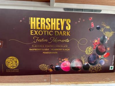 The image shows a close-up of a Hershey's Exotic Dark Festive Moments chocolate box. The box is a deep maroon color with gold lettering and intricate gold line drawings of various berries and pomegranates. The text on the box reads "HERSHEY'S EXOTIC DARK Festive Moments FLAVORED CENTER CHOCOLATE RASPBERRY & GOJI | BLUEBERRY & AÇAI POMEGRANATE". There is also a QR code with the text "SCAN TO SURPRISE YOUR LOVED ONES". The box is resting on a wooden surface with visible screws. The lighting is bright, suggesting an indoor setting during the daytime. The overall presentation is festive and inviting, hinting at a holiday or special occasion gift. The location context of San Jose del Monte, Philippines, does not directly influence the visual description of the image, but it provides geographical context for the product's potential availability or origin. The image focuses solely on the product packaging, with no people or broader scene visible.