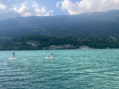 Two individuals are paddleboarding on the turquoise waters of Lake Brienz, near Bönigen, Switzerland, under a partly cloudy sky. Both subjects are oriented with their backs to the camera, wearing light-colored hats and blue personal flotation devices. The person on the left stands upright on a white paddleboard, holding a black paddle. The person on the right is kneeling or sitting on a white paddleboard, also holding a paddle, and wears pink shorts or swim trunks. The water surface shows moderate ripples. The background features heavily forested mountains rising abruptly from the far shoreline. A prominent light-colored, horizontal geological formation is visible on the lower mountain slope. Scattered buildings with reddish roofs are discernible amidst the trees on the distant shoreline. The sky exhibits patches of blue interspersed with white and grey cumulus clouds.