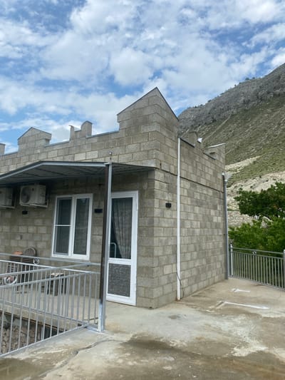 In Chalda, Russia, a two-story building constructed of gray concrete blocks features a crenellated rooftop design. The building has a metal awning over a section of its facade, beneath which two air conditioning units are mounted. A balcony or terrace in front of the building is enclosed by a silver metal railing. Two large windows and a glass door with white frames are visible on the lower level. In the background, a steep, rocky mountain slope is covered with sparse vegetation. The sky is partly cloudy, with white clouds against a blue backdrop. The overall scene depicts an outdoor view of a building in a mountainous region.