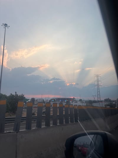 The image displays an exterior scene captured from a vehicle on an elevated roadway, likely an overpass or bridge, during sunset. The location is identified as Tultitlán, Mexico, specifically near the Jorobas interchange on a major highway.

The upper two-thirds of the frame are dominated by a sky with scattered clouds, through which prominent crepuscular rays of sunlight are visible, fanning out from a point obscured by the clouds. The lower horizon, particularly towards the center-left, exhibits a distinct pinkish-orange glow.

In the immediate foreground, the bottom right corner shows the reflection within a vehicle's side mirror, depicting a queue of stationary or slow-moving traffic, primarily consisting of red passenger automobiles. A concrete barrier extends horizontally across the lower third of the image, featuring approximately twelve grey vertical support posts, each capped with a yellow band.

Below and beyond this barrier, a multi-lane highway or expressway is discernible. On the far left, a tall street light pole equipped with multiple lamp fixtures rises vertically. To its right, a green overhead highway sign is partially visible, indicating "Jorobas." Further along the highway, structures consistent with toll booths or a highway interchange are visible, featuring multiple illuminated signals displaying green and red lights. On the right side of the frame, a large metal electricity transmission tower stands prominently, with a smaller similar pylon visible in the distance behind it. Various distant structures, possibly urban or industrial, are discernible along the horizon.