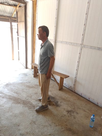 In Olomoda, Nigeria, an adult male stands in profile, facing left, within an interior space. He is wearing a light blue-grey short-sleeved polo shirt, light beige trousers, and grey athletic shoes. He has short dark hair and wears glasses. His hands are positioned at his sides. The floor is rough concrete with visible dirt and irregular dark stains. To the right, a wall is composed of multiple light-colored panels framed by white strips, creating a grid pattern. Two simple wooden benches with angled supports are placed against this panelled wall behind the man. To the left, a brightly lit open doorway or passageway reveals an exterior view, including a corrugated metal roof section and wooden structural elements. A blue plastic water bottle with a white cap is visible on the concrete floor in the lower right foreground. No direct actions or interactions are depicted.