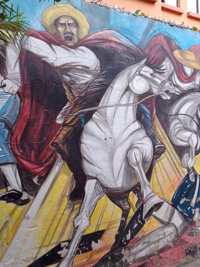 A mural adorns a wall in Municipio Tarija, Bolivia. The artwork depicts a scene of figures on horseback, possibly representing historical or revolutionary figures. One rider, wearing a yellow sombrero and a red cape, is shown mid-yell with his mouth open. The horse is white. A figure on the left of the image is in partial view. Another horse and rider are visible on the right side. The background is a mix of light blue, yellow, and other colors, with what appears to be sunlight emanating from the figures. A row of broken glass bottles sits along the top edge of the wall to prevent people from climbing over.