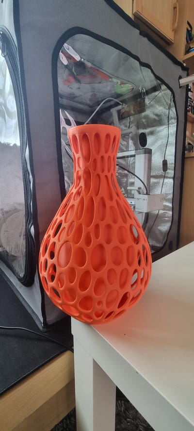 An orange, 3D-printed vase with a lattice-like pattern of holes sits on a white table. The vase has a rounded base that tapers towards a narrow neck. Behind the vase is a grey fabric tent with a clear plastic window. Inside the tent, some items are visible, including what appears to be a white 3D printer. To the right of the tent, a wooden cabinet is partially visible. The image is taken from a slightly elevated angle, and the lighting suggests an indoor setting. The location is not discernible from the image itself, and therefore cannot be specified as being in Naaldwijk, Netherlands.