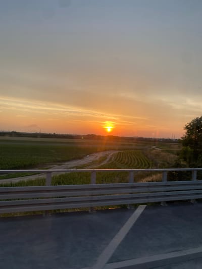 The image captures a sunset scene over farmland in Podłęże, Poland. The sun, a bright orange orb, is low on the horizon, casting a warm glow across the sky and illuminating wispy clouds. Below the sky, a vast expanse of agricultural fields stretches out, with rows of crops and a dirt path winding through them. In the distance, industrial structures and smokestacks are visible, suggesting the presence of industry near the rural landscape. A highway guardrail runs across the foreground, with a road surface visible below it. The overall atmosphere is serene, with the vibrant colors of the sunset creating a picturesque view.