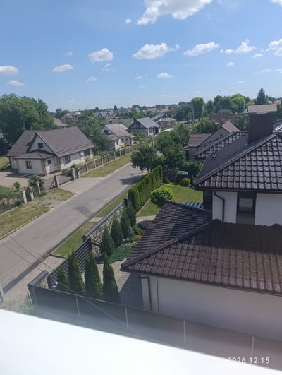 The image captures a suburban residential scene under a bright, partly cloudy sky. Several houses with dark tiled roofs are visible, nestled amongst lush green trees. The architecture appears to be modern, with a mix of light-colored facades and dark trim. A paved road runs through the neighborhood, bordered by fences and well-maintained gardens with manicured hedges and small evergreen trees. In the foreground, a large house with a dark roof is prominent, suggesting a recent construction or renovation. The scene is bathed in sunlight, indicating it is daytime, likely mid-morning or afternoon. The overall impression is one of a peaceful, well-kept community. A timestamp in the bottom right corner reads "26.06.2026 12:15", indicating the date and time the photo was taken.