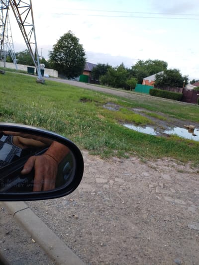 The image depicts a view from inside a vehicle, showing a reflection in the side mirror of a person's arm and hand resting on the car door. The background is a rural or semi-rural scene in Taganrog City District, Russia. A tall metal structure, likely a power line pylon, is visible on the left. Beyond a grassy area with some puddles, there are houses with green and orange accents, as well as fences and trees, suggesting a residential area. The weather appears to be overcast or nearing dusk, with a pale sky. The ground is a mixture of gravel and dirt, with some patches of grass. The text "TS IN MIRROR ARE CLOSER THAN THEY APPEAR" is visible in the lower portion of the side mirror. The overall impression is that of a casual drive through a moderately developed area.