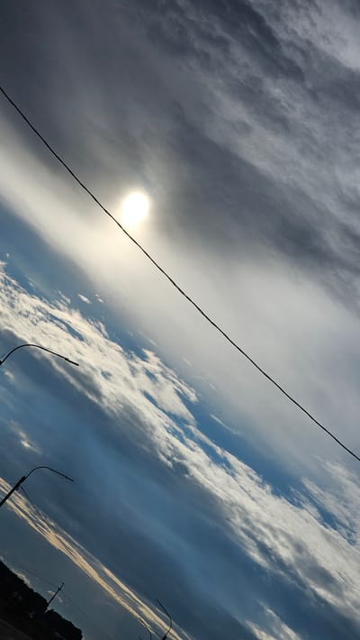The image shows a sky with a bright sun peeking through scattered clouds. The sun is a luminous orb in the upper left quadrant of the frame. The clouds are a mix of white and grey, some appearing wispy and others more dense, creating a textured sky. A dark, thin wire stretches diagonally across the entire frame from the upper left to the lower right. In the lower portion of the image, two streetlights with curved poles are visible. The image is tilted, giving the impression of looking upwards at an angle. The lower edge of the frame shows a dark silhouetted landscape, possibly trees or buildings, and a few more power lines. The lighting suggests it is daytime, possibly late afternoon or early morning given the angle of the sun and the mix of light and shadow in the clouds. There are no people or specific landmarks identifiable within the frame to pinpoint the exact location within Покровское, Russia, however, the presence of streetlights and power lines suggests an urban or suburban environment.