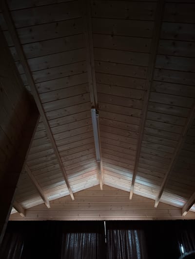 The image shows a wooden plank ceiling with visible rafters, in a room that appears to be dimly lit by a fluorescent light fixture suspended from the ceiling. Dark curtains are visible at the bottom of the frame. The architecture suggests a rustic or unfinished interior, possibly a barn, shed, or a room with a vaulted ceiling. The location is specified as Machara, Abkhazia. There are no people or any discernible actions taking place.
