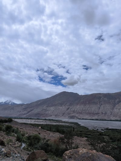 This is a wide-angle, landscape photograph taken in Panamik, India, under a cloudy sky. The foreground features rugged terrain with scattered rocks and sparse vegetation. A small stream or body of water is visible to the left, winding through the rocky landscape. In the midground, a wide, dry riverbed or floodplain stretches across the scene, flanked by dense green vegetation on either side. Further in the distance, majestic mountains rise, their slopes layered with different shades of rock and earth. The peaks of some of the distant mountains are capped with snow, contrasting with the earthy tones of the lower slopes. The sky is filled with a dramatic array of clouds, ranging from light and wispy to darker, more ominous formations, with patches of blue sky visible through the gaps. The overall impression is of a vast, natural landscape in a remote, mountainous region.