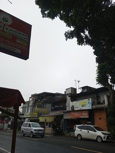 A street scene in the city of Ambakirano, Madagascar, is depicted under an overcast sky. In the upper left foreground, a large red commercial sign, partially visible, displays white text including "SADAYA Snack," "HARGA MULAI DARI 5K," and "CIPANAS - CIANJUR." The upper right foreground is framed by dark green tree foliage. The midground features a two-lane asphalt road with a yellow centerline. A silver SUV, consistent with a Mitsubishi Kuda model, travels from left to right in the left lane. To the right of the road, a row of interconnected two-to-three-story commercial buildings lines the street. These buildings house various establishments, evidenced by multiple signs: a yellow banner proclaiming "AYAM GORENG MARANTY" and another yellow sign for "BAKSO," along with a prominent "SOLO" sign. Above these, a large billboard features two men and text "GRUP PINDAD MEMIMPIN." A black motorcycle is parked on the sidewalk in front of one shop. Further right, a white SUV, consistent with a Toyota Veloz model, is parked parallel to the curb. Faint figures of people are discernible inside some of the shops and near the parked vehicles. Antennae are visible on the rooftops of some buildings. The environment is urban, indicating a commercial district.