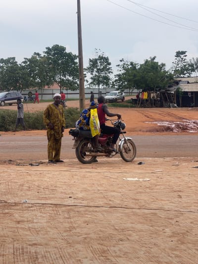 In Osu, Nigeria, a scene unfolds on a dusty, unpaved road. Two men are the central figures. One man, dressed in a traditional yellow and brown patterned outfit and a white cap, stands on the left side of the frame, facing the camera with his hands at his sides. To his right, a second man is seated on a red motorcycle, wearing a red sleeveless shirt and black pants. He is facing towards the right, with the motorcycle's engine appearing to be running. Another individual, partially obscured by the rider, is seated behind him on the motorcycle, wearing a blue and yellow patterned outfit and holding a bright yellow bag. The motorcycle is equipped with a luggage rack. In the background, a row of trees lines the horizon, and a few simple structures with corrugated metal roofs are visible. A few vehicles, including a white SUV and a darker colored car, are parked further back. The ground is a reddish-brown dirt, with some muddy patches and scattered debris. The sky is overcast.
