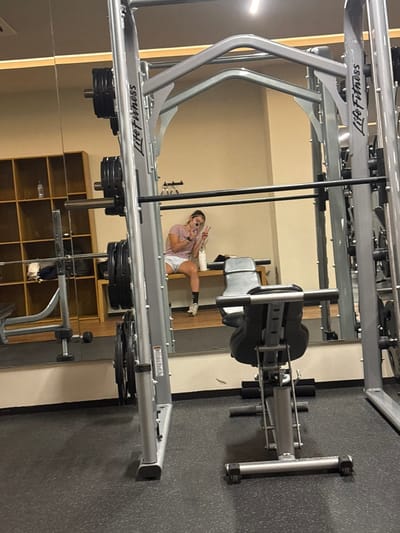 The image shows a mirrored gym setting with exercise equipment, including a Life Fitness machine with weight plates. A woman is reflected in the mirror, posing with a peace sign while holding a phone, presumably taking a selfie. Background details include storage shelves, wall paneling, and rubber flooring. The image is rotated, causing disorientation.