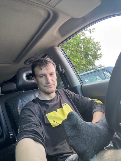 This is a first-person perspective, slightly rotated, selfie taken inside a vehicle. The subject is a man with short, dark hair and stubble, looking directly at the camera with a neutral expression. He is wearing a black long-sleeved shirt with a yellow graphic on the chest. His right leg is extended, and he is wearing a dark grey sock. The interior of the car is visible, including a grey headliner and a black leather seat. Through the side window and sunroof, green trees and a bright, overcast sky are visible, suggesting it is daytime. The scene appears to be in Shatava, Ukraine, based on the provided location context. There is no discernible text in the image.