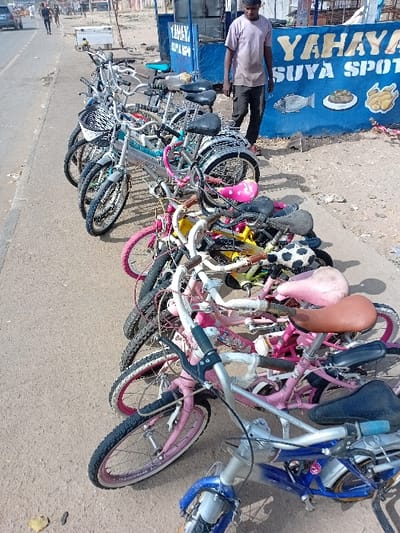 The image depicts an outdoor scene in Sharada, Nigeria, at a roadside location under bright daylight. Approximately two dozen bicycles, ranging from children's sizes to adult models, are clustered on unpaved ground adjacent to a paved road. The bicycles display various colors including pink, blue, silver, and black, with different seat styles and handlebar configurations.
An adult male, wearing a light-colored t-shirt and dark trousers, stands to the right, positioned near a blue commercial stall. The stall is labeled "YAHAYA SUYA SPOT" in white and yellow text, featuring painted representations of a fish, a plate of food, and a chicken.
The paved road extends into the background, where indistinct human figures, vehicles, and structures are visible. The ground cover in the immediate foreground consists of paved asphalt, transitioning to dry, unpaved earth where the bicycles are arranged. A single yellow leaf rests on the paved surface in the lower left. No direct interactions between individuals or with the bicycles are observed.