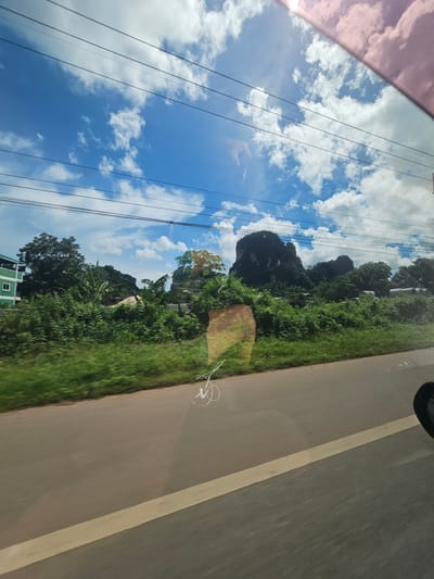 This image captures a scenic view from a moving vehicle in Ban Thung, Thailand. The foreground is dominated by the vehicle's windshield, which reflects the bright blue sky dotted with fluffy white clouds. Sunlight, tinged with pink from the vehicle's tinting, streaks across the top left of the frame, suggesting it's daytime. Several power lines crisscross the sky, indicating the presence of infrastructure.

The scene outside the vehicle is a lush, green landscape. Dense foliage, including trees and what appear to be banana plants, lines the side of a paved road. A prominent, dark, rocky outcrop, partially covered in vegetation, rises dramatically from the greenery in the middle ground. A small, green building with a white-framed window is visible in the bottom left corner, adding a touch of human habitation to the natural scenery. The road itself has a light-colored line marking its center, and the texture of the asphalt suggests it's a well-traveled route. A faint reflection of what looks like a pair of white earbuds is visible on the road surface, a subtle detail hinting at the activity within the vehicle. The overall impression is one of a bright, sunny day with a sense of movement, likely during travel through a rural or semi-rural area.