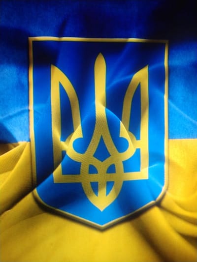 The image displays the national emblem of Ukraine, the Tryzub, superimposed on the Ukrainian flag. The emblem, rendered in yellow, is centrally positioned on a blue background, which is the upper half of the flag. The lower half of the flag is yellow. The fabric appears to be textured and is draped, creating folds and shadows that add depth to the image. The lighting highlights the texture of the fabric and the edges of the emblem. The image is a close-up shot, focusing on the flag and its emblem. There are no people or environmental elements visible, nor is there any indication of its specific location within Vylok, Ukraine.