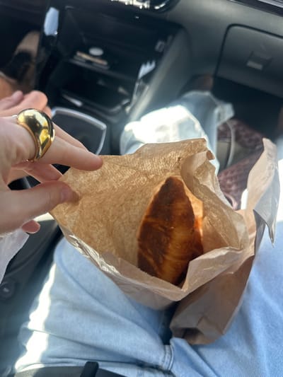 The scene depicts the interior of a vehicle, located in Mafra, Portugal. A person's left hand, adorned with a prominent, rounded gold-colored ring on the ring finger, holds an open brown paper bag. Inside the bag, a golden-brown pastry, consistent with a croissant or similar laminated dough, is partially visible. The hand is positioned over the lap of another individual, whose legs are clad in light blue denim pants. The vehicle's dark-colored dashboard and center console are visible in the background, exhibiting compartments and controls. Natural daylight illuminates the scene, casting highlights and shadows within the cabin.
