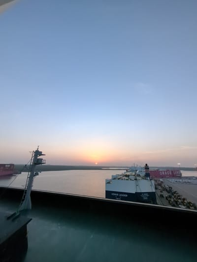 The image shows a sunrise over the port of Hambantota, Sri Lanka. The sun is low on the horizon, casting a warm glow over the scene. In the foreground, the deck of a ship is visible, with a mast and some equipment. In the middle ground, the water of the port is calm, reflecting the colors of the sunrise. Several ships are docked in the port, including a large cargo ship with the name "VENUS LEADER" and "TOKYO" visible, and another vessel with "GRACE LINE" written on its side. The background shows a green, vegetated shoreline and distant structures. The sky is a clear, pale blue, with the sun creating an orange and pink gradient near the horizon. The scene is peaceful and serene, capturing the early morning light at the port.