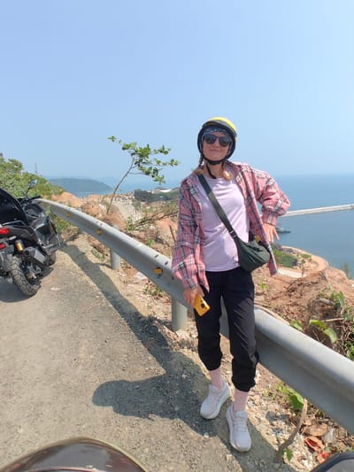 An adult female subject stands on the paved shoulder of a road, overlooking a coastal vista in Hải Vân Ward, Vietnam. The subject, positioned centrally, wears a yellow open-face helmet over a blue patterned bandana, dark sunglasses, a light purple t-shirt beneath an open pink and white plaid long-sleeve shirt, black cuffed pants, light pink socks, and white athletic shoes. A dark green cross-body bag is slung over her left shoulder. She holds a yellow smartphone vertically in her right hand, while her left hand rests on her hip. To her left, partially visible, is a parked dark grey adventure-style motorcycle. A silver metal guardrail with yellow circular reflectors spans horizontally across the mid-ground. Beyond the guardrail, a barren, rocky hillside slopes down to a developed coastal area featuring buildings, roads, and a concrete breakwater extending into the calm blue ocean. The sky above is clear and light blue. The scene is illuminated by bright sunlight, casting visible shadows.