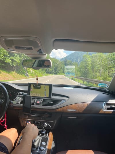 The image shows the interior of a car driving on a road in Oberammergau, Germany. The viewpoint is from the passenger seat, looking forward. The dashboard is visible, with a navigation screen displaying a map and route information. A hand is on the gear shift. The exterior view through the windshield shows a paved road, trees, and mountains in the distance. There are also signs visible on the side of the road. The lighting suggests it is daytime.