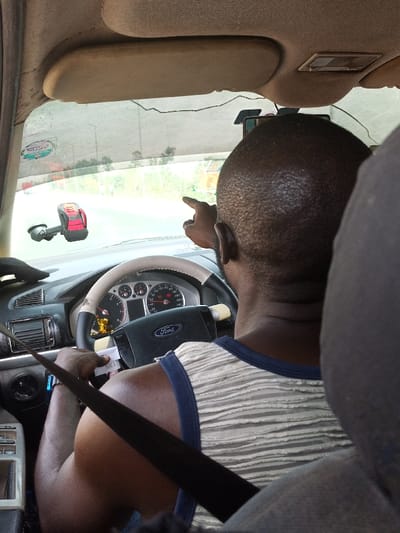 The image captures a view from the back seat of a car, looking forward at the driver and the road ahead in Kubare, Nigeria.
The primary subject is the driver, seen from behind. He appears to be a man with short, dark hair and dark skin, wearing a white and blue patterned sleeveless top (tank top). A seatbelt is fastened across his chest and shoulder. His right arm is extended, with his index finger pointing forward, seemingly indicating something on the road or in the distance. His left hand is resting on the steering wheel, holding a small, white piece of paper or ticket.
The car's interior is visible, featuring a dashboard with analog gauges, and a steering wheel with a "Ford" logo at its center and a beige cover over its rim. A device, possibly a phone holder, is suction-cupped to the windshield on the left. The car's headliner and sun visors appear to be light-colored and somewhat worn.
Through the windshield, a clear, paved road is visible, flanked by green foliage and trees on both sides, suggesting a rural or suburban setting. The bright lighting indicates it is daytime. No specific weather conditions like rain or heavy clouds are apparent.
Visible text includes the word "Ford" on the steering wheel. Other text on a sticker on the windshield and on the paper held by the driver is too small and blurry to be deciphered.