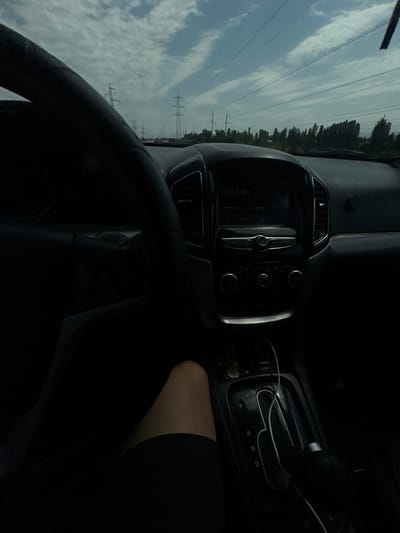 The image depicts the interior of a car, likely a Jeep Grand Cherokee, viewed from the driver's perspective. The dashboard and steering wheel are prominently featured, with a digital display showing "101.0 MHZ." Outside the car, a cloudy sky and a line of power transmission towers are visible, suggesting a suburban or rural setting. A section of the driver's leg, clad in dark shorts, is also visible in the foreground. The car's gear shift is in the "P" position. The overall lighting suggests it is daytime, but the interior of the car is dimly lit. The car is likely parked or stopped, as evidenced by the gear selection and the visible leg position. No other individuals are apparent in the vehicle. The location is specified as Istiqlol, Uzbekistan.