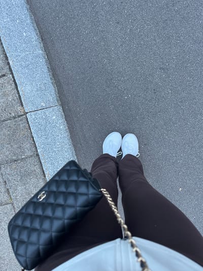 A close-up, top-down view captures the lower body of an individual standing on dark grey asphalt. The individual wears dark brown trousers and white athletic shoes. A black quilted handbag, featuring a gold-colored interlocking C logo (consistent with Chanel) and a gold-colored chain strap, is positioned on the left side. A light-colored garment with a zipper is visible on the upper thigh. To the immediate left of the asphalt, a light grey stone curb separates the pavement from a sidewalk composed of light grey paver stones. Sparse dried vegetation is visible in the cracks of the paver stones. The scene represents an outdoor urban or suburban setting during daylight. The precise location within Skanderborg, Denmark, is not discernible from the visual evidence.