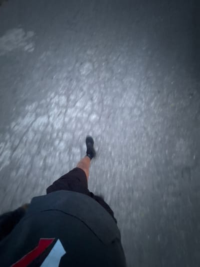 The image shows a person walking on what appears to be a paved surface. The person is wearing black shoes, black shorts, and a black top with a red and white design. The ground appears to be a gray or light-colored paved surface. The image is taken from a high angle, looking down at the person's legs and feet. The location is not specified.