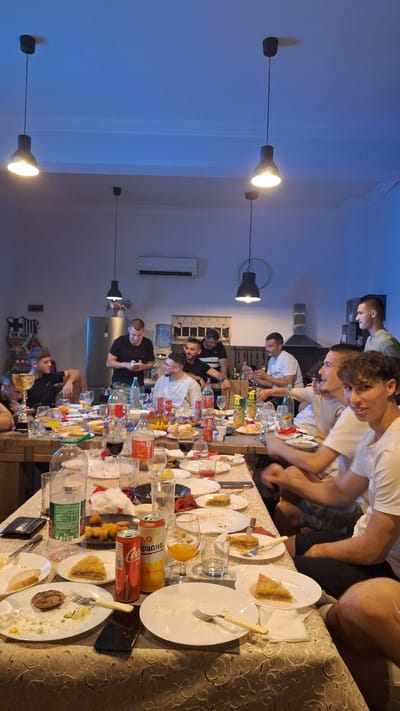A group of people are sitting around a long table covered with dishes, glasses, and beverages, seemingly after a meal. They are interacting and socializing. The location is Gorski Izvor, Bulgaria. The room has an air conditioner and hanging light fixtures.