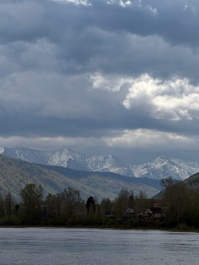 The image depicts a view of mountainous terrain near the city of Сизая, Russia. Snow-capped mountains are visible in the center of the frame, contrasted against a cloudy sky to the left. A body of water, possibly a lake or river, is on the right. The land near the water's edge has trees, houses, and utility poles.