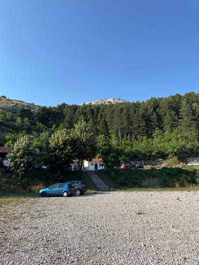 The image depicts a gravel parking area in front of a wooded hillside and a clear blue sky. In the foreground, two cars are parked on the gravel: a blue hatchback and a dark-colored SUV. Behind them, a small white building with a red roof is visible, with stairs leading up to its entrance. Further up the hill, a dense forest of pine trees covers the landscape, leading to a rocky, mountainous peak in the distance. Several other vehicles, including a red pickup truck and a white van, are parked along a road higher up on the hillside. The scene appears to be a remote location, possibly in a rural area like Bukovica, Montenegro, given the natural surroundings and the presence of a small dwelling and parking area.