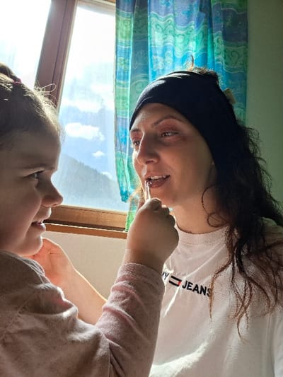 The image captures an indoor scene in Davos, Switzerland, illuminated by natural light from a window. Two individuals, an adult woman and a young child, are positioned next to the window.

On the left, a young girl, estimated to be between three and six years old, is shown in profile, facing right. She has dark hair partially tied back and wears a light pink long-sleeved top. Her left arm is raised, and her hand holds a clear tube applicator, which she is actively placing against the adult woman's lips. Her attention is focused on the adult.

On the right, an adult woman, appearing to be in her mid-20s to late 30s, is seen with her head turned slightly left, facing the child. She has dark, wavy hair, mostly contained by a black headband. She wears a white short-sleeved t-shirt featuring the "TOMMY JEANS" logo in black and red text across the chest. Her mouth is slightly open, indicating she is receiving the application to her lips. Pink eyeshadow is visible on her eyelids.

The background features a wooden-framed window, through which a bright sky with white clouds is visible above a steep, tree-covered mountainous landscape, consistent with an alpine environment. To the right of the window, a curtain with a blue and green patterned design is visible. The immediate interior wall appears light-colored.