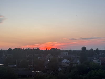 The image depicts a rural landscape in the village of Pratasewickaje, Belarus, at dusk. The sky is a gradient of pale blue at the top, transitioning to soft orange and vibrant red near the horizon, indicating sunset. A thin band of pink and orange clouds stretches horizontally across the sky. The lower half of the image is dominated by a dense canopy of dark green trees, interspersed with the rooftops of houses. The houses appear to be of various sizes and styles, with some featuring dark, sloped roofs and others with lighter colored facades. Some houses are partially obscured by the foliage. A few tall, slender structures, possibly utility poles or small towers, are visible rising above the treeline in the distance. The overall impression is one of a tranquil, pastoral scene as day transitions into night.