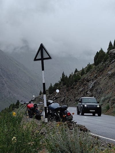 The image depicts a cloudy, overcast day in the mountainous terrain of Darcha, India. A dark-colored SUV, possibly a Land Rover Defender, is driving on a wet asphalt road that curves to the right. The road is bordered by a steep, rocky mountainside dotted with sparse coniferous trees. In the foreground, two motorcycles are parked on the shoulder of the road, which is covered in gravel and vegetation. A triangular road sign with black and white markings is mounted on a pole between the motorcycles. The red motorcycle has a license plate that reads "HP66 A4765". The grey motorcycle is partially obscured by the red one and vegetation. The background shows more rugged, mist-covered mountains.