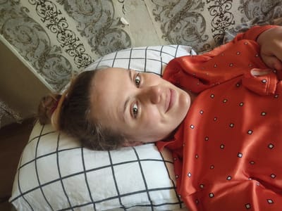 A young woman with blonde hair tied back is lying on a pillow with a black and white checkered pattern. She is looking directly at the camera with a slight smile. She is wearing a bright orange top with a repeating pattern of small, dark diamonds. The background appears to be a wall with ornate, vintage-style wallpaper featuring a damask or floral motif in muted tones of grey and brown. The lighting suggests it could be daytime, possibly indoors. The overall mood is relaxed and casual. There is no visible text in the image. The location is identified as a suburban settlement in Ukraine.