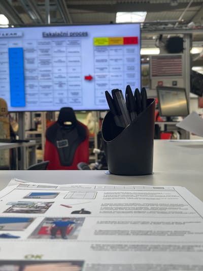 The image shows a close-up view of a desk with various items. In the foreground, a black pen holder is filled with pens and markers. To the right of the pen holder, a document with text and diagrams is laid out. The document appears to be instructional, possibly a manual or guide, with several small images depicting actions or objects.
Behind the desk, a large screen displays a grid of information, with some cells highlighted in red and yellow. The text on the screen is partially visible, with "Eskalační proces" (Escalation process) clearly legible on the left side.
Further in the background, a red and black seat, resembling a racing car seat, is visible. The setting appears to be an office or workshop environment, with shelves and equipment in the distance. The lighting suggests it is daytime. There are no people clearly visible in the frame, but the items suggest an active workspace. The overall impression is one of a functional, organized work area.