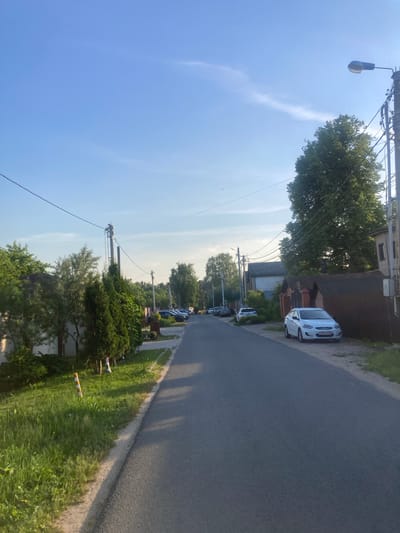 The image depicts a residential street scene in Novoaleksandrovo, Russia, captured under a clear, light blue sky with faint, wispy clouds visible in the upper right quadrant. The sun, positioned out of frame to the upper right, casts long shadows across the asphalt road, indicating late afternoon or early evening.
The asphalt road, dark grey and without markings, extends from the foreground into the background, narrowing slightly.
On the left side of the road, a strip of unpaved ground with green grass and some dirt runs parallel to the asphalt. A small, vertically oriented pole with alternating orange and white stripes is visible in this grassy area in the foreground. Behind this strip, a variety of vegetation, including manicured evergreen hedges and deciduous trees, lines the street. Utility poles with multiple overhead wires are present along this side, receding into the distance. Portions of residential fences and structures are discernible through the foliage.
On the right side of the road, a strip of dirt and sparse grass separates the pavement from a prominent brown fence constructed of vertical wooden slats. A white four-door Hyundai Accent sedan is parked on this dirt strip, parallel to the fence, facing away from the camera. The vehicle's rear license plate displays "X 903 TO 190". Further down the street, multiple other vehicles are parked. A tall utility pole, heavily laden with numerous black wires, stands near the fence in the mid-ground, extending upwards into the sky. A blue-housed streetlight fixture is attached to this pole. Beyond the fence and utility pole, a large, dense deciduous tree with green foliage dominates the right mid-ground, and parts of additional residential buildings are visible.
No individuals or active traffic are visible in the scene. The environment appears to be a quiet suburban or semi-rural residential area.