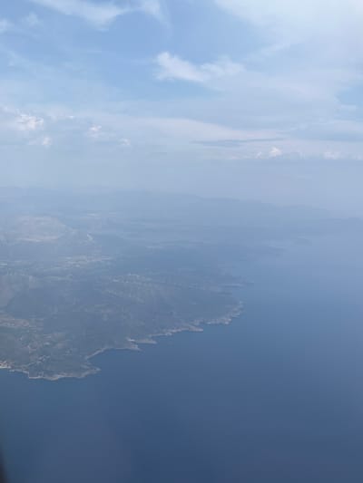 An aerial or high-altitude view captures a vast, hazy landscape under a pale blue sky, depicting a rugged coastline meeting the sea. The image, when oriented correctly, shows a significant body of dark blue water on the left, merging with a prominent, rocky landmass on the right.
The landmass appears mountainous and arid, characterized by sharp, jagged rock formations and slopes. Scattered across these slopes, particularly in the mid-ground and right side of the image, are numerous man-made structures that resemble terracing, fences, or pathways. These appear as white or light-colored lines and segments, suggesting human modification of the steep terrain, possibly for agriculture, infrastructure, or access. The land recedes into a distant, heavily obscured background due to atmospheric haze.
The sky, occupying the upper left and blending into the hazy atmosphere over the land, is a light blue with scattered, wispy white clouds. The overall weather appears hazy or misty, softening the distant features and diffusing the sunlight, suggesting daytime. No specific human activity or event is visible, but the man-made structures imply a human presence and interaction with the environment. There is no visible text in the image. Given the location context, this scene likely depicts a coastal area within the Spata-Loutsa Municipal Unit in Greece.