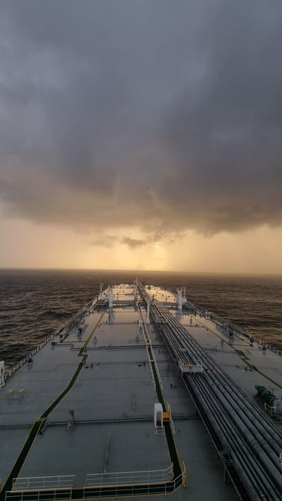 An aerial perspective shot captures the deck of a large cargo ship sailing on a choppy, dark sea under a stormy, overcast sky. The ship's deck is vast and mostly gray, with green and yellow markings delineating pathways. A prominent cluster of large pipes runs along the right side of the deck, stretching towards the horizon. In the distance, the sea meets the sky, with a bright, hazy band of sunlight breaking through the heavy clouds, casting a golden glow on the horizon and creating a dramatic, atmospheric effect. The rough texture of the sea suggests strong winds. There are no visible people or signs of human activity on the deck.