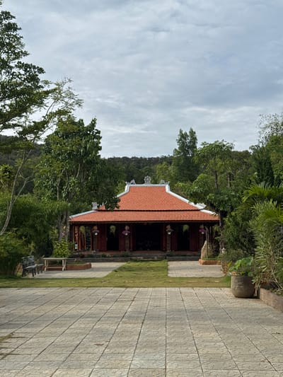 The image shows a traditional Vietnamese temple in Liên Nghĩa, Vietnam. The temple has a red-tiled roof, dark wooden pillars, and hanging flower baskets. It is surrounded by lush green trees and vegetation. A paved walkway leads to the temple's entrance. The sky is overcast. A bench is placed near the temple entrance.