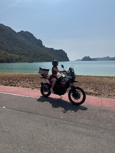 A motorcycle with a rider wearing a helmet is parked on the edge of a road. A side case is attached to the motorcycle. The road is adjacent to a shoreline with tropical islands in the background. The sky is clear and blue.